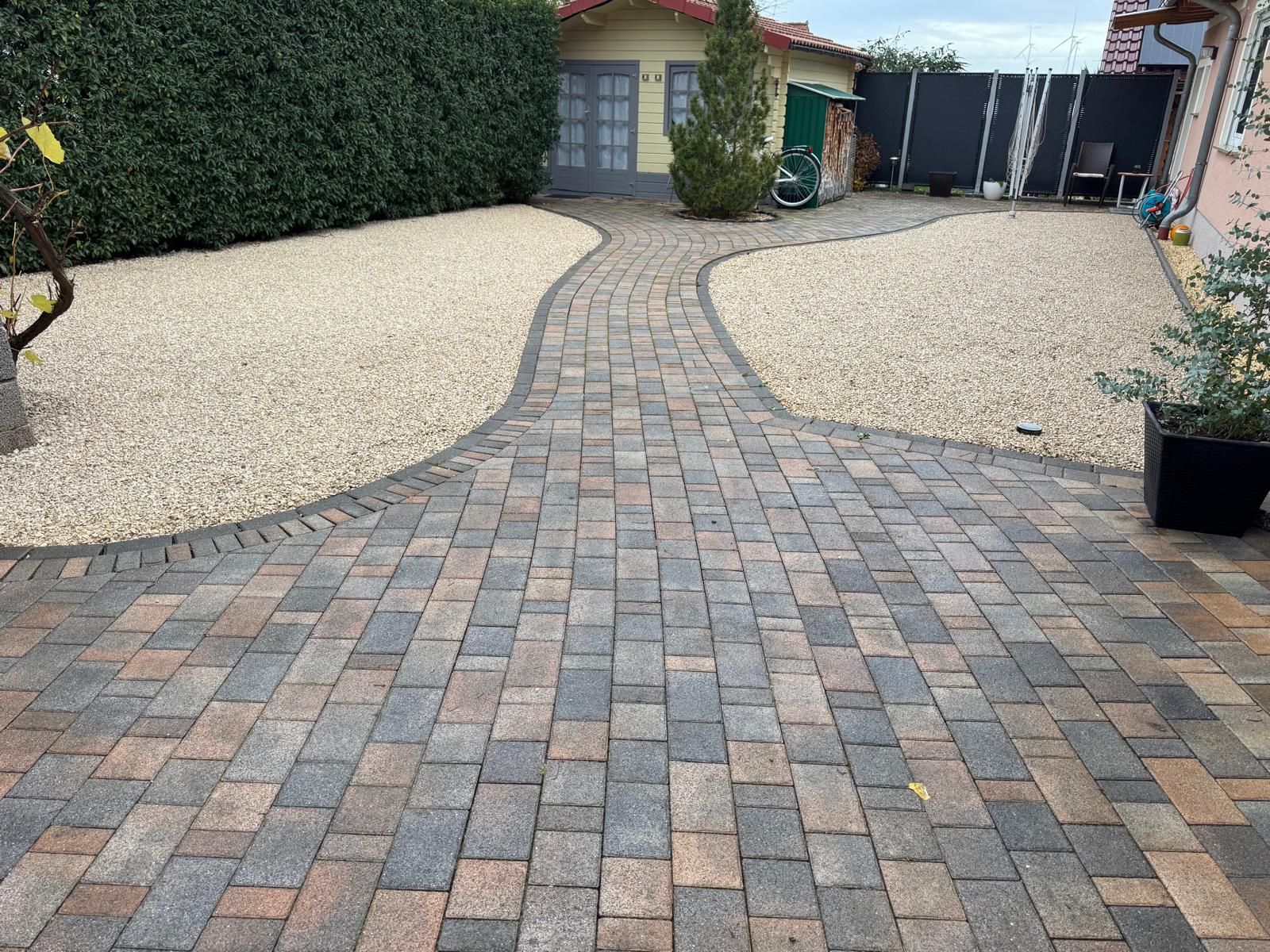 Ein Ziegelweg führt zu einem Kieshof mit einem kleinen Schuppen und Grünpflanzen.