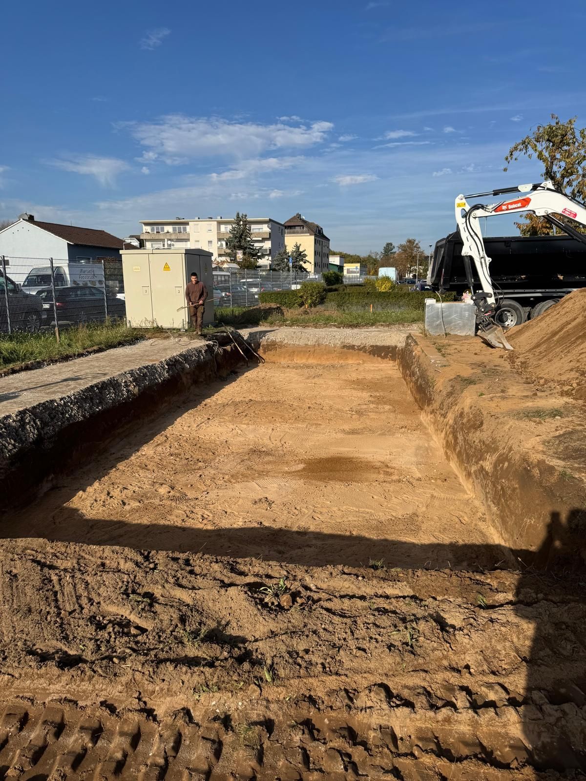 Aushubstelle mit einem rechteckigen Loch. Ein Bagger und Verteilerkästen heben sich von Gebäuden und blauem Himmel ab.
