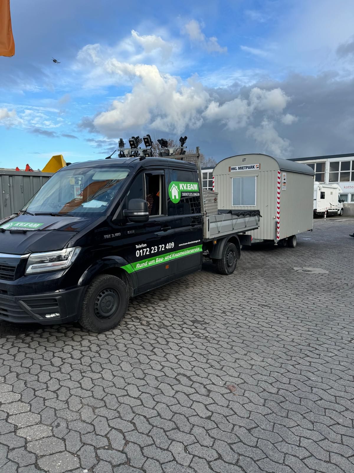 Schwarzer Lieferwagen mit kleinem Anhänger auf gepflasterter Fläche. Bewölkter Himmel im Hintergrund.