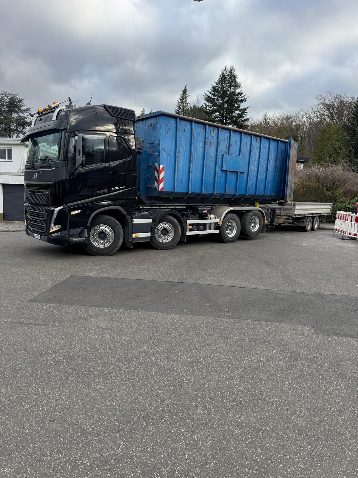 Schwarzer Sattelzug mit blauem Container auf einem gepflasterten Parkplatz; Anhänger im Schlepptau; bedeckter Himmel.