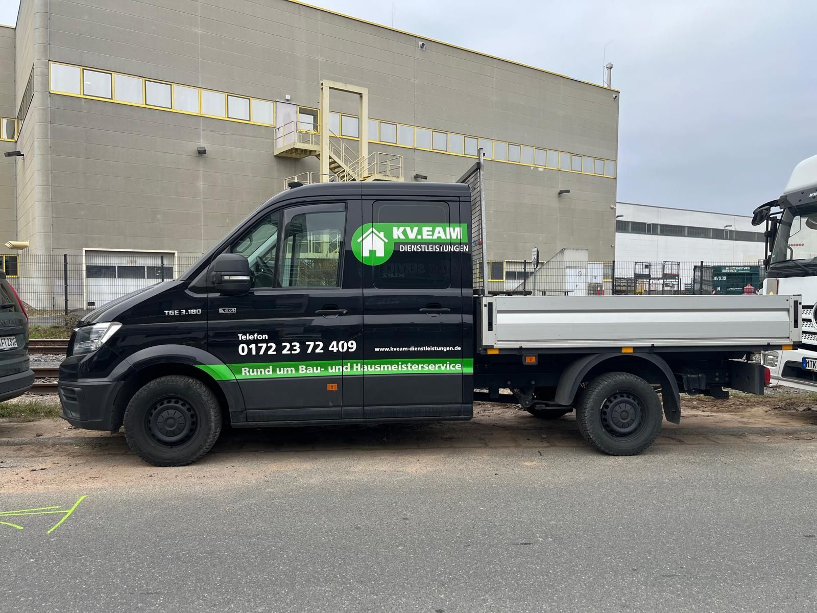 Ein schwarzer KV.EAN-LKW mit Pritsche parkt vor einem grauen Gebäude.
