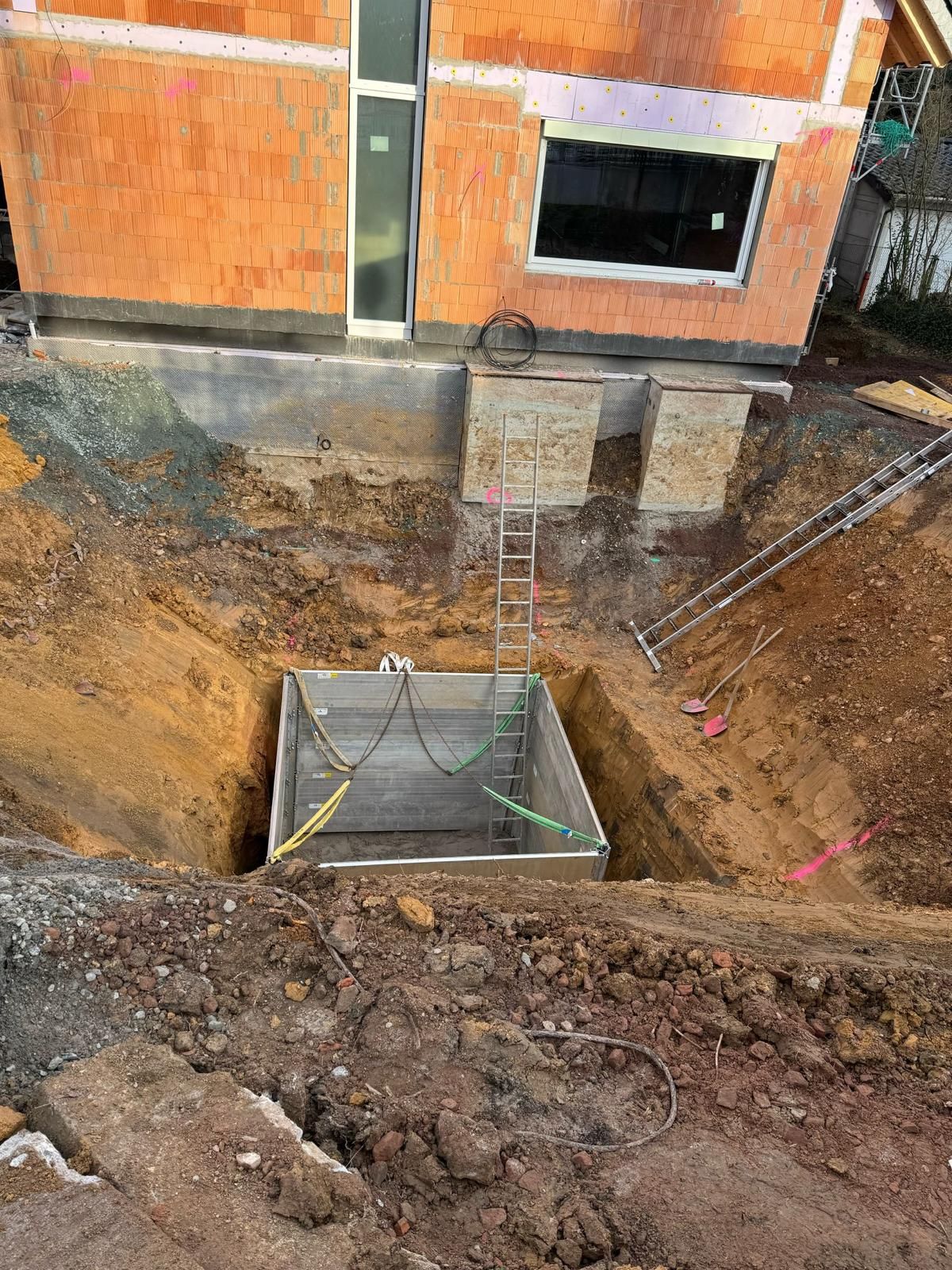 Baustelle mit ausgehobener Baugrube. Eine Betonkonstruktion wird neben einem Backsteingebäude in die Baugrube abgesenkt.