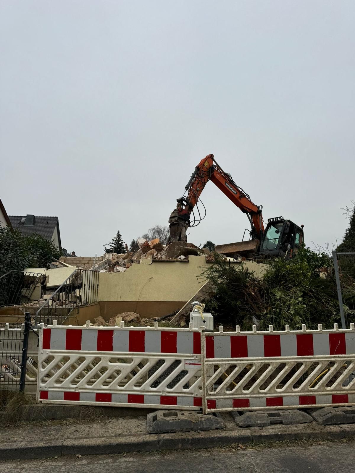 Ein Bagger reißt ein Gebäude hinter einer rot-weißen Absperrung ab; bedeckter Himmel.