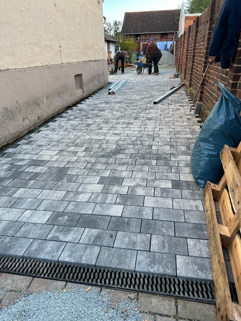 Arbeiter pflastern eine schmale Gasse mit grauen Steinblöcken, entlang einer Backsteinmauer und eines Gebäudes.