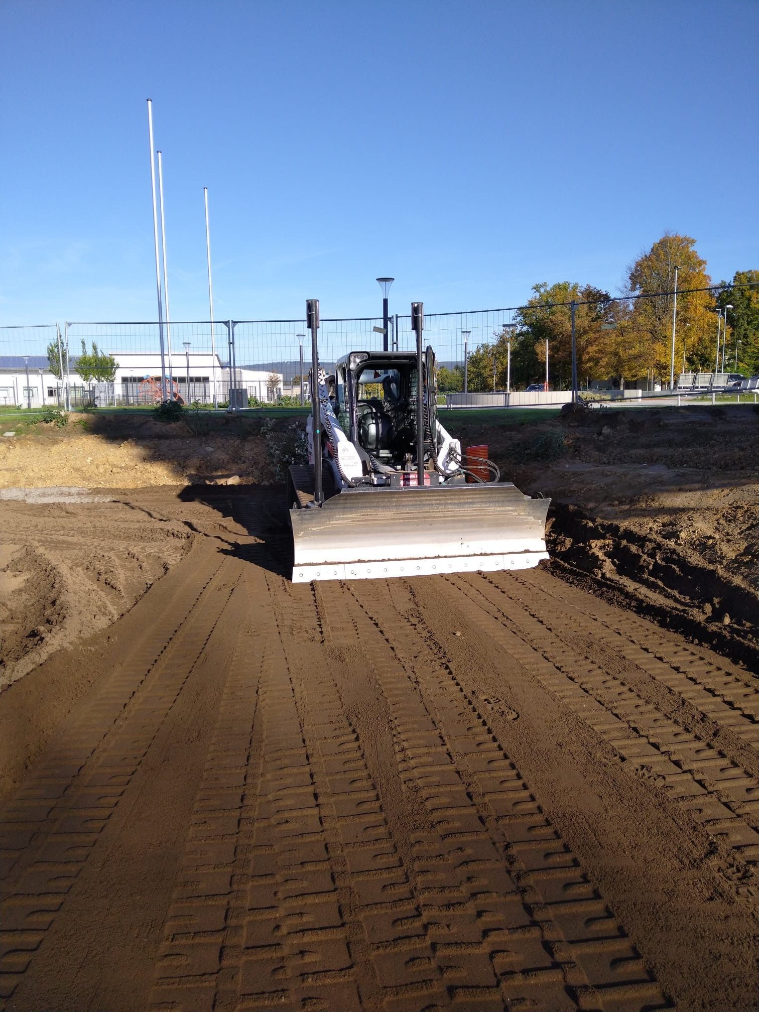 Ein Kompaktlader mit Graderschild ebnet unter blauem Himmel braunen Boden auf einer Baustelle.