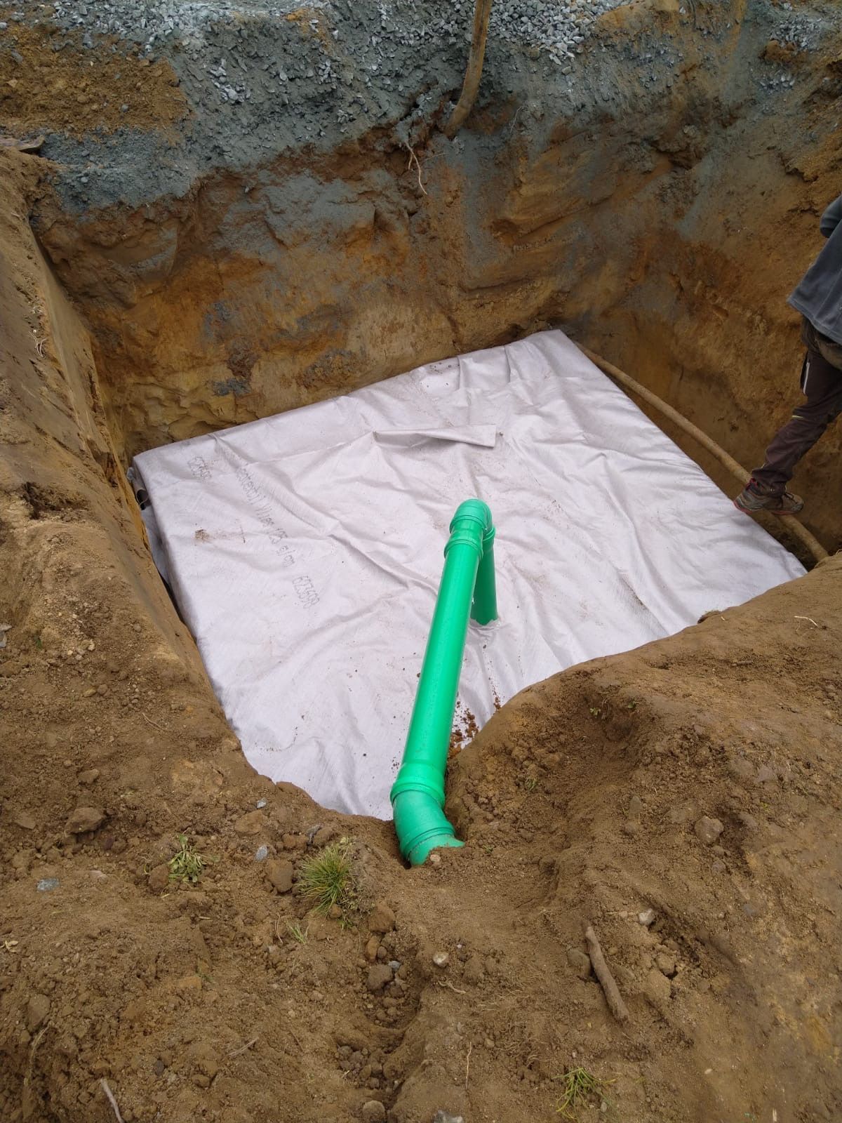 Ein quadratisches Loch im Boden mit einer weißen Plane und einem grünen Rohr darin. Im Hintergrund ist jemand zu sehen.