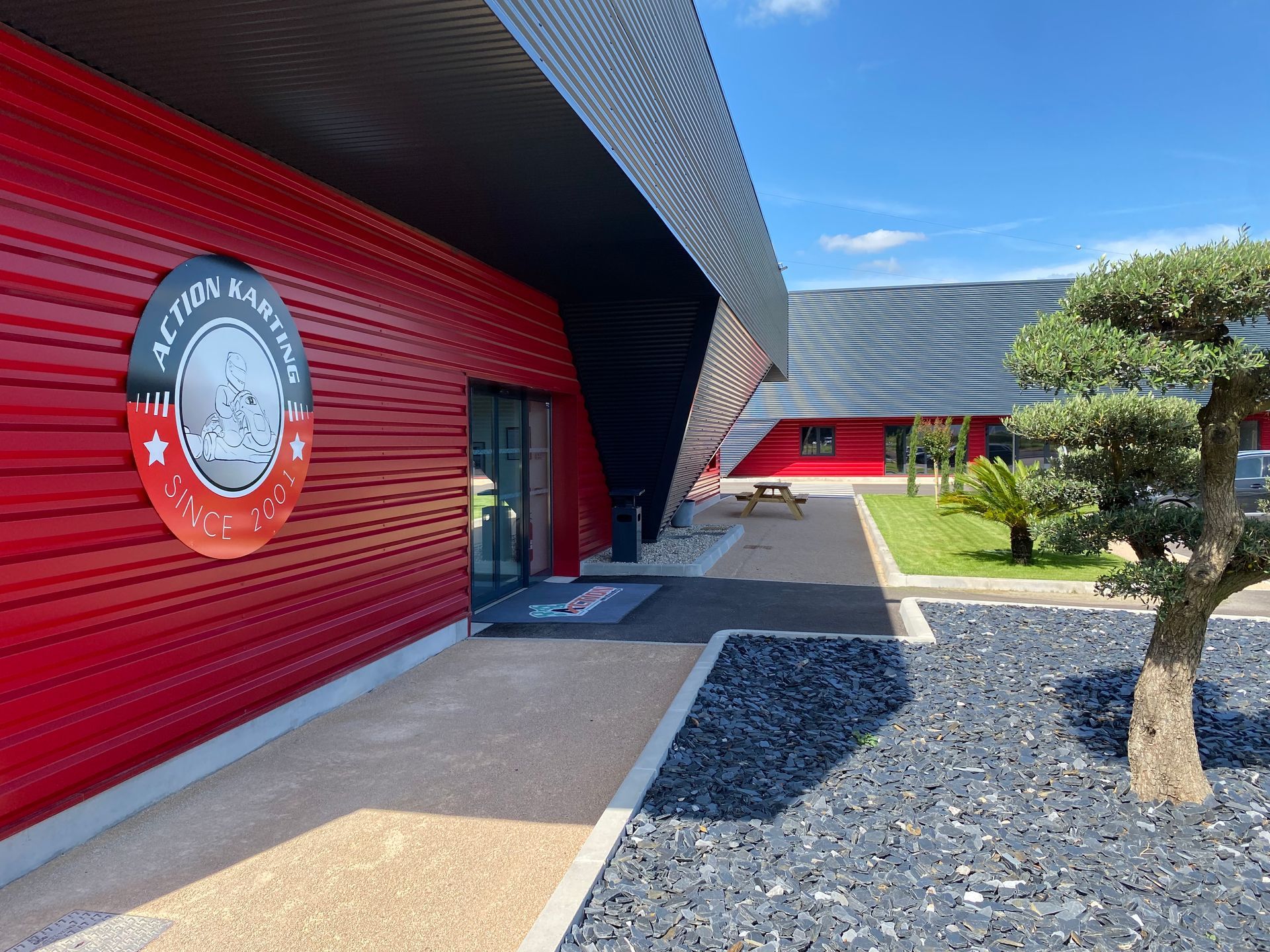 Bâtiment rouge avec panneau « Action Karting », toit noir, chemin en pierre et un bonsaï.
