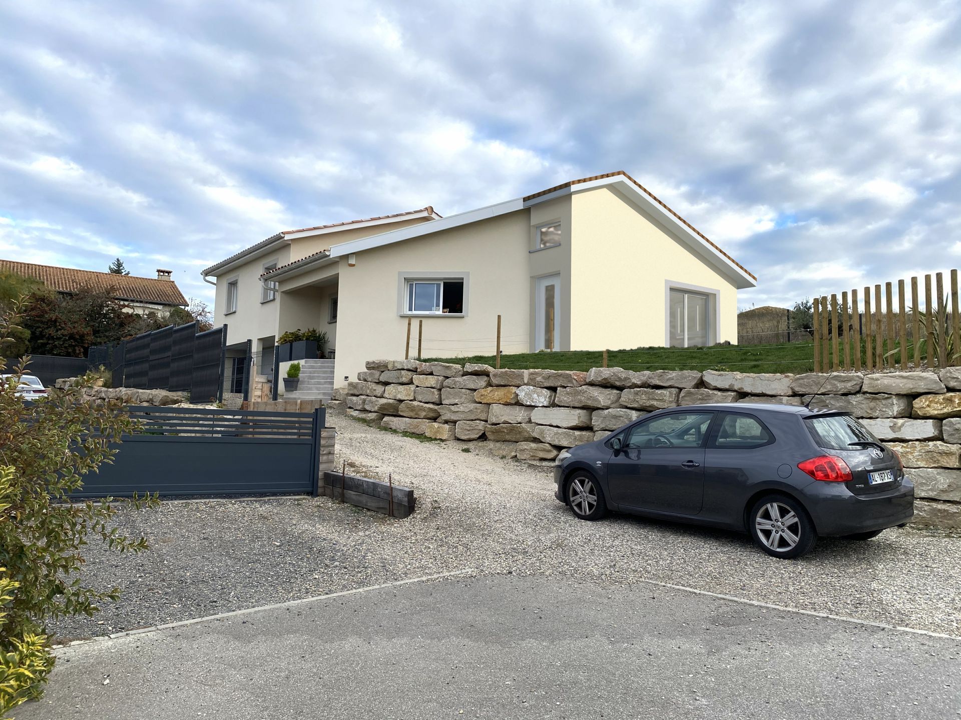 Voiture grise garée devant une maison beige clair avec une allée de gravier sous un ciel nuageux.
