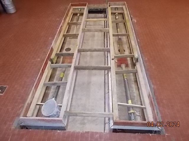 Eine rechteckige Bodenöffnung mit Holzrahmen, vermutlich für Maschinen oder Wartungsarbeiten. Rötlicher Boden.