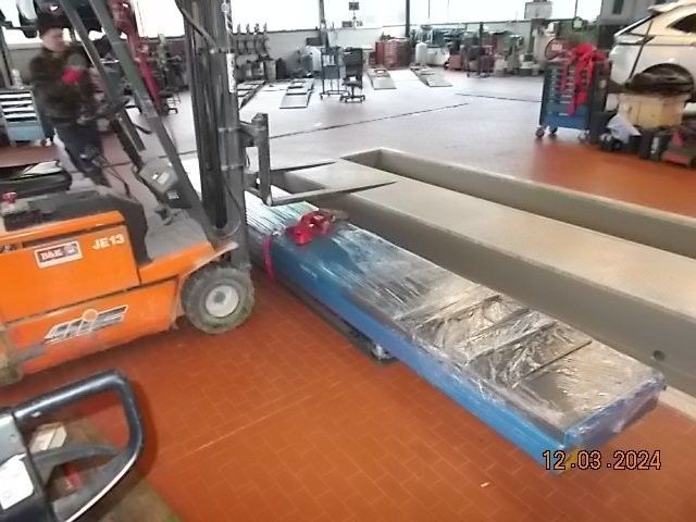 Ein Gabelstapler steht in einer Autowerkstatt neben einem blau verpackten Gegenstand. Orange, graue und rote Farben sind zu erkennen.