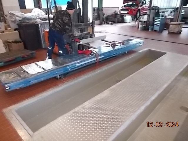 Ein Mechaniker arbeitet in einer Werkstattgrube und hält Werkzeug in der Hand. Im Hintergrund ist ein rotes Auto zu sehen.