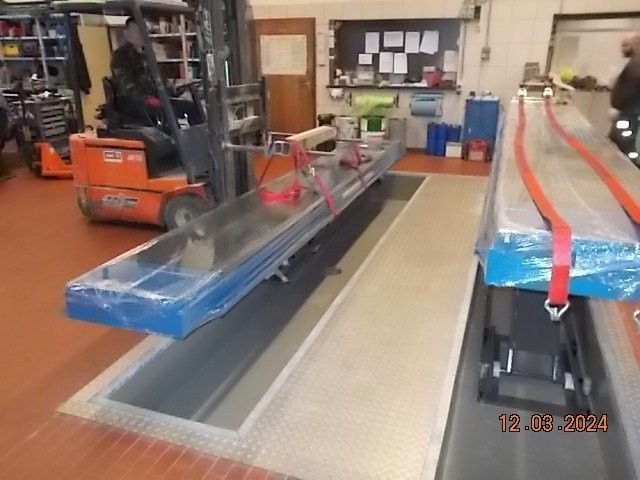 Zwei Hebebühnen in einer Garage. Eine ist mit Gurten gesichert und blau umwickelt; im Hintergrund ein orangefarbener Gabelstapler.