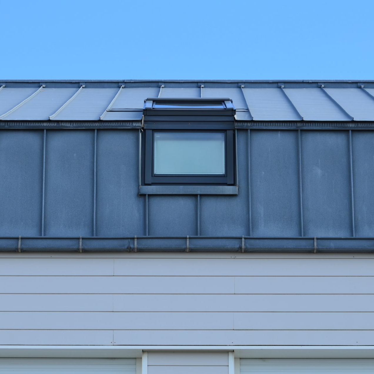 Toit bleu-gris d'un bâtiment avec une petite lucarne et un bardage métallique.