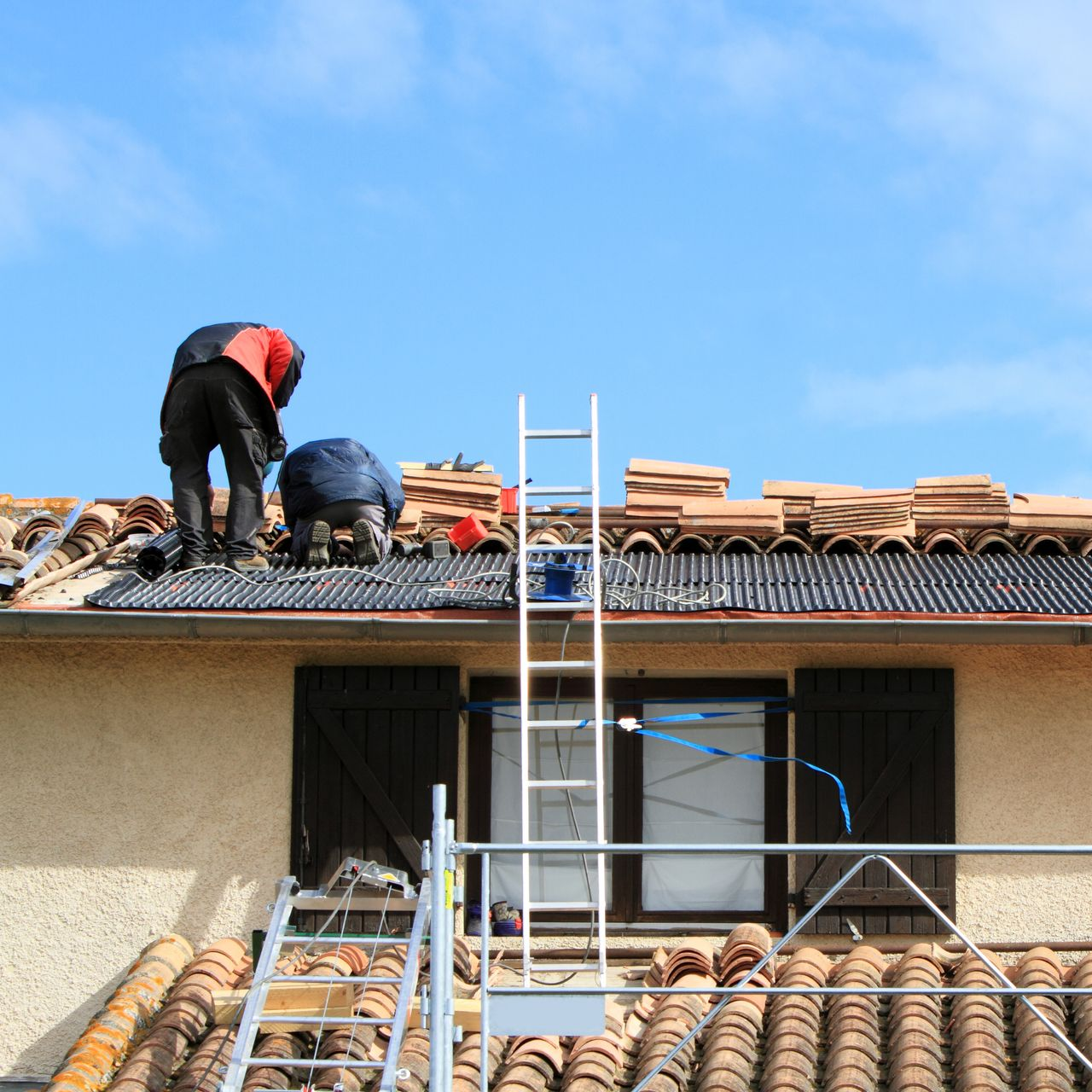 Des ouvriers réparent le toit d'une maison avec des tuiles et des échelles.