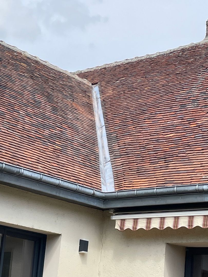 Toit de tuiles rouges avec cheminée et gouttière blanches sur un bâtiment beige sous un ciel bleu.