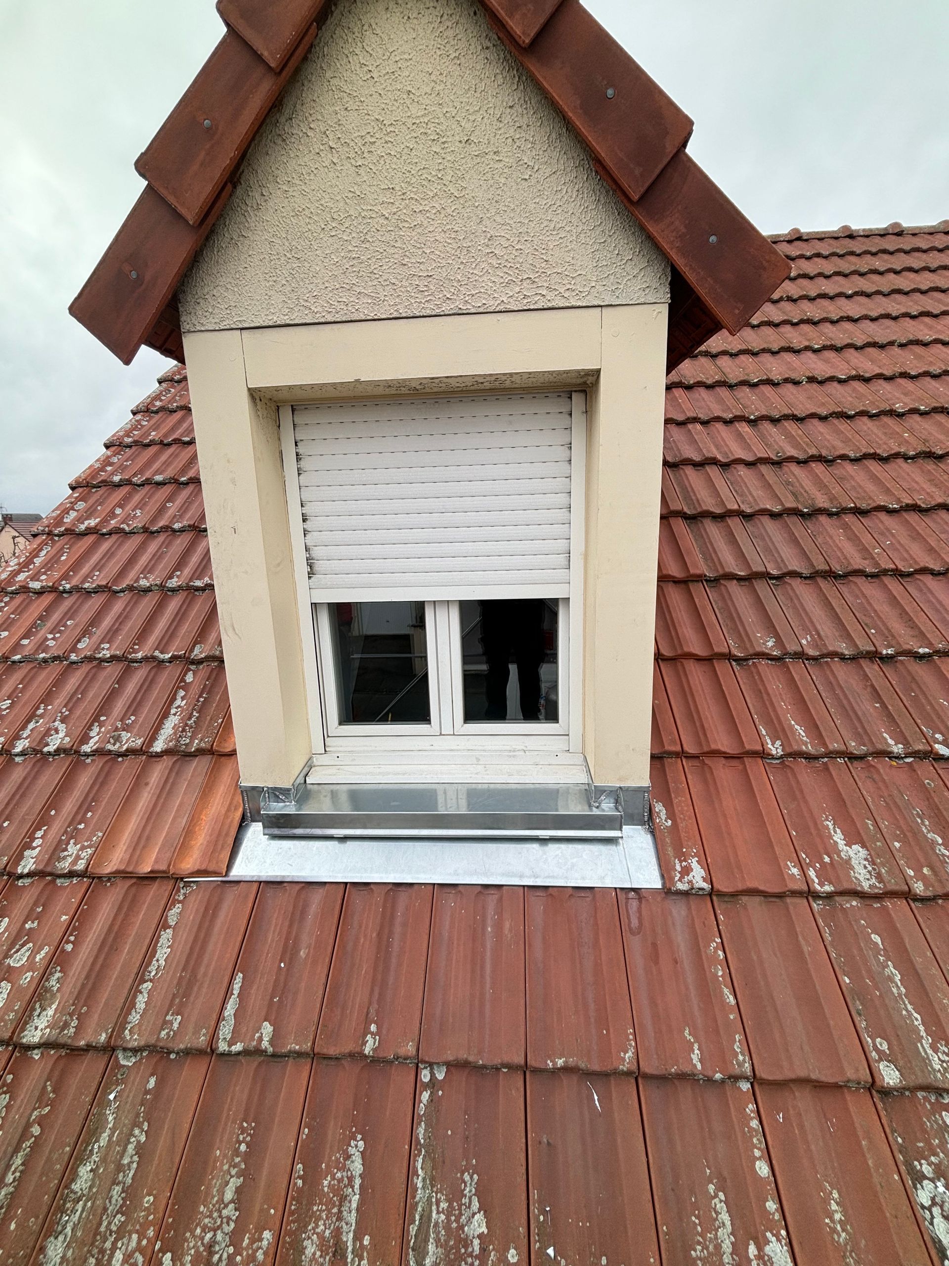 Lucarne sous un toit de tuiles rouges avec un volet blanc partiellement ouvert.