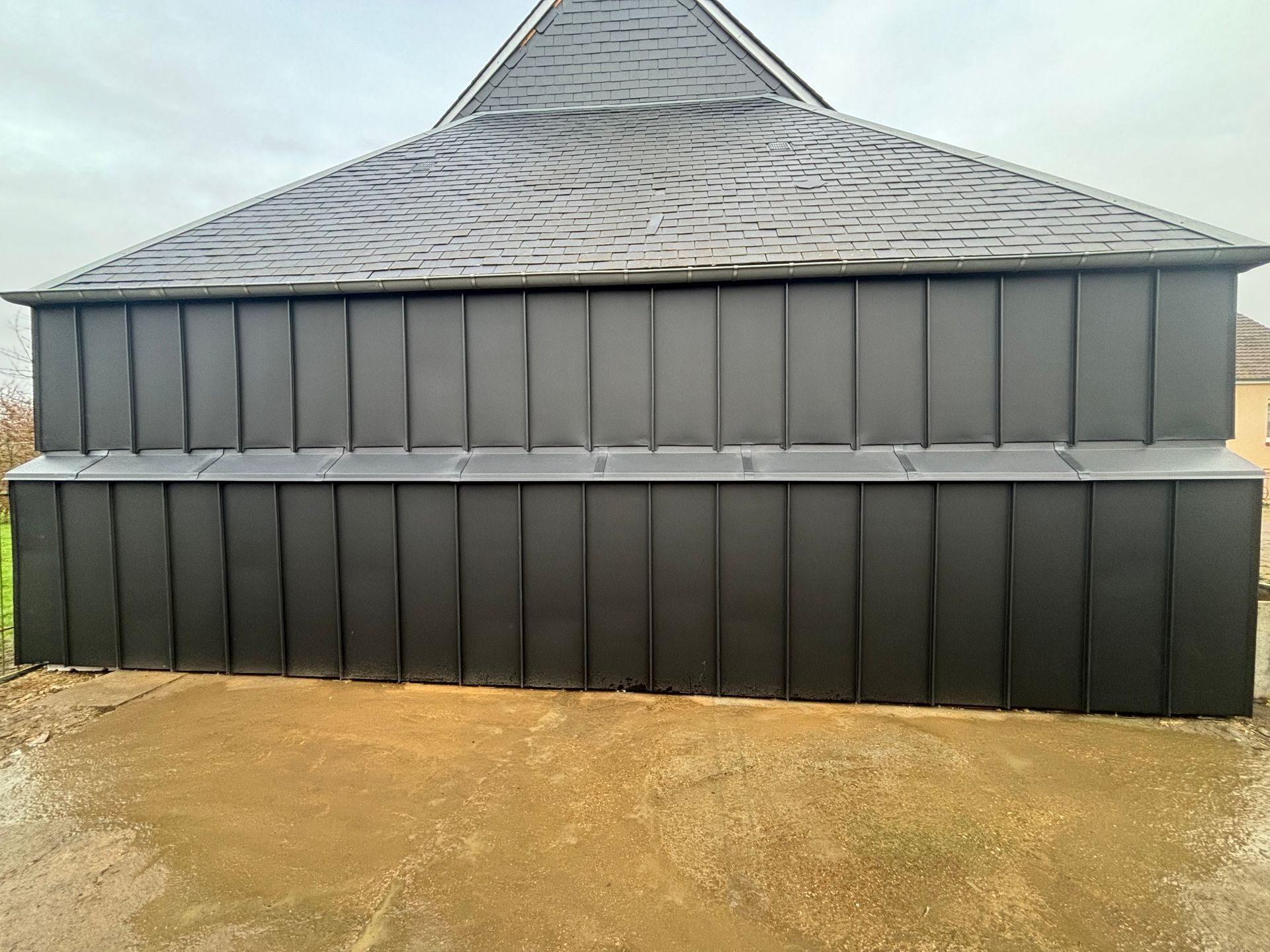 Bâtiment de deux étages à revêtement métallique gris et toit conique en bardeaux, construit sur un terrain nu.