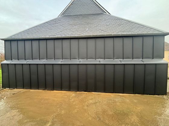 Un bâtiment au toit en métal noir, avec un bardage supérieur gris et un bardage inférieur noir, sur un terrain en terre battue.