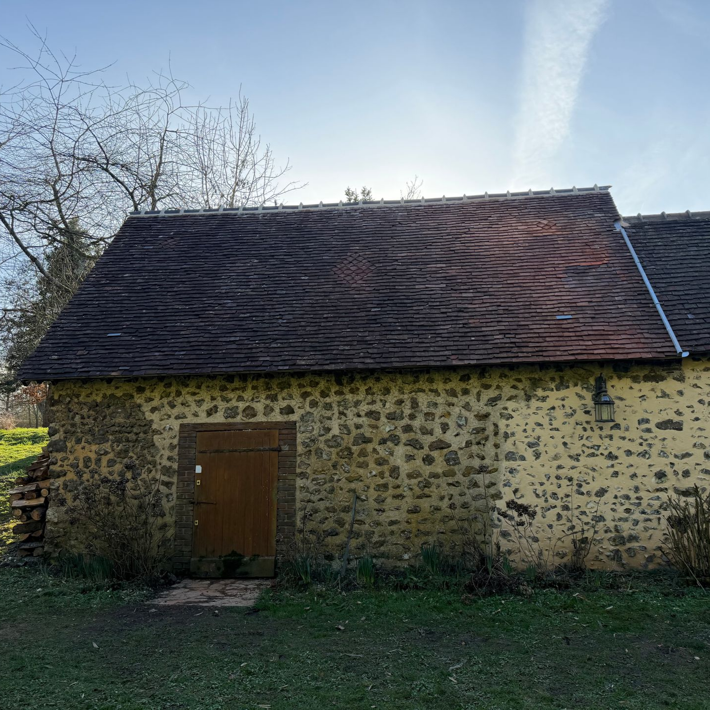 Dépendance en pierre avec un toit de tuiles sombres, une porte en bois et une cour herbeuse sous un ciel lumineux