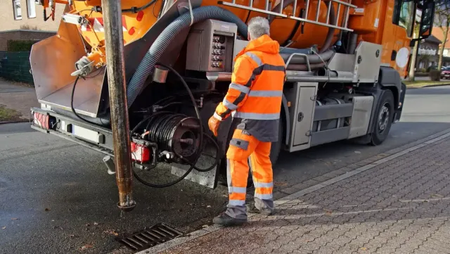 Arbeiter im orangefarbenen Anzug neben einem LKW, der einen Abwasserkanal auf einer Kopfsteinpflasterstraße wartet.