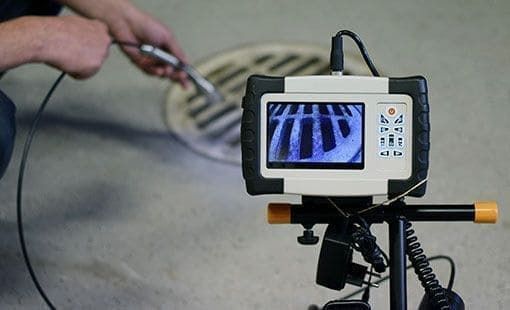 Person, die mit einer Kanalinspektionskamera in einen Abfluss schaut. Auf dem Kamerabildschirm ist ein Gitter zu sehen.