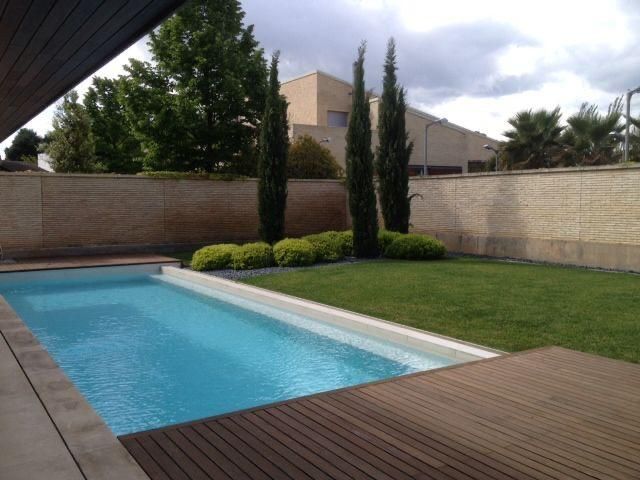Patio trasero con una estrecha piscina azul, terraza de madera, césped y altos cipreses bajo un cielo nublado.