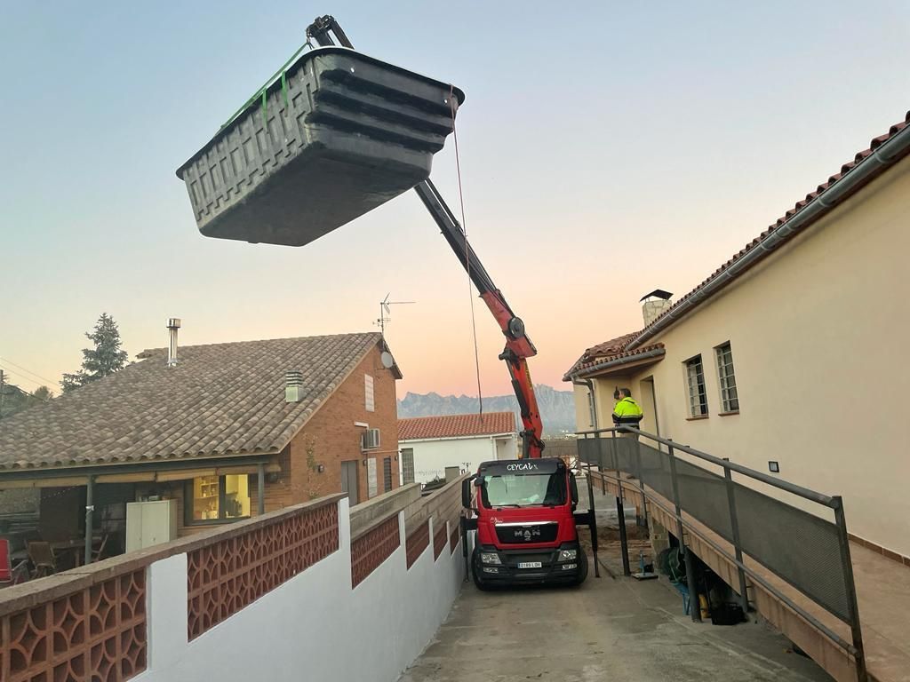 Una grúa eleva un gran contenedor sobre una estrecha calle residencial junto a las casas al atardecer.
