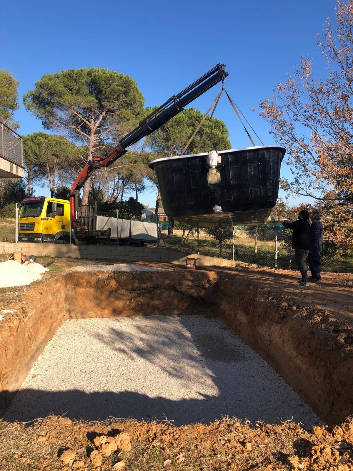 Una grúa eleva una pala negra sobre una gran excavación con grava y trabajadores al aire libre.