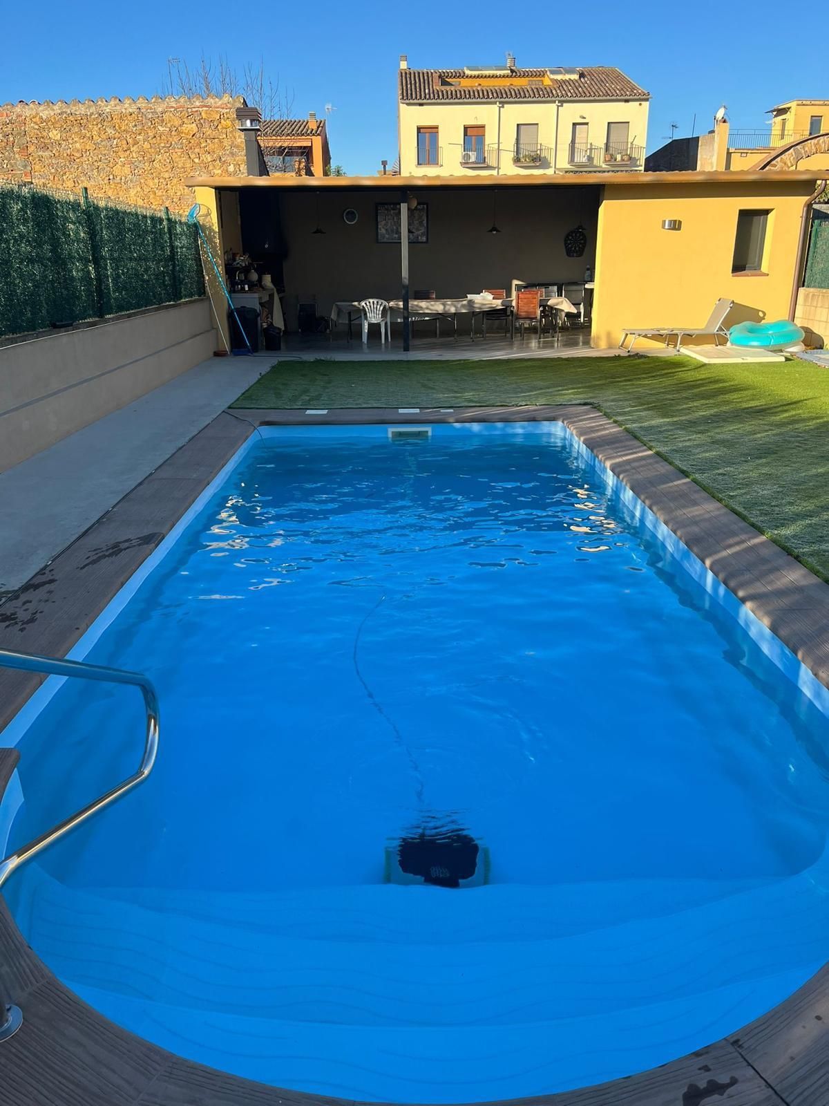 Piscina en el patio trasero con cubierta azul, asientos en el patio y una casa amarilla al fondo.