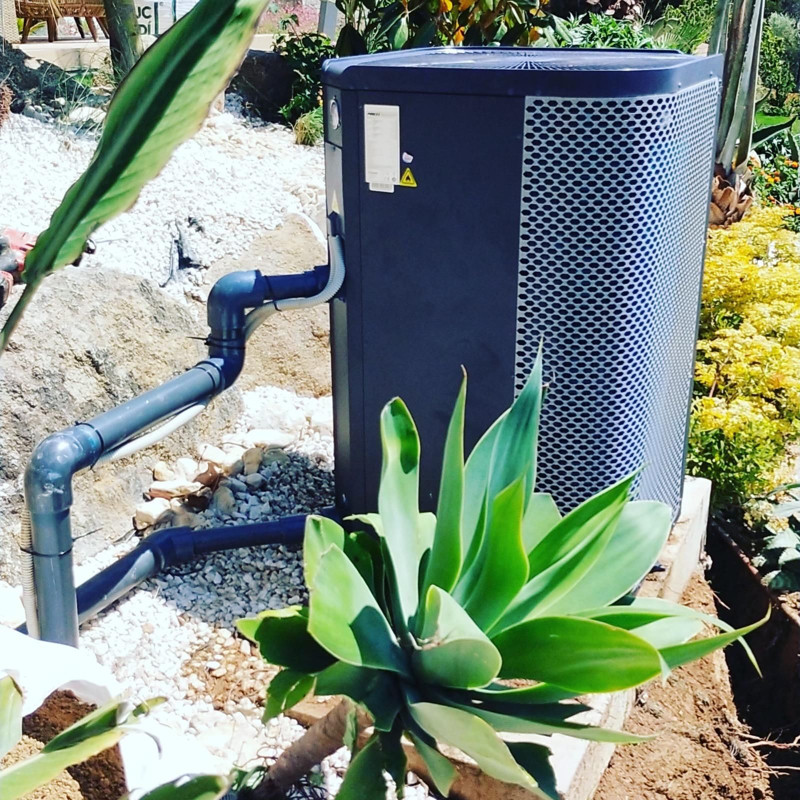 Unidad de aire acondicionado exterior situada junto a rocas y plantas tropicales, con tuberías negras conectadas.
