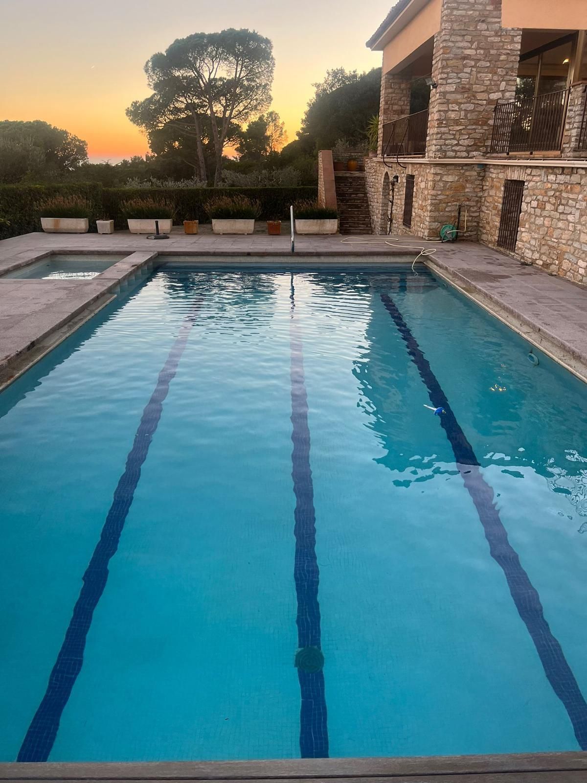 Piscina rectangular junto a una casa de ladrillo al atardecer, con tumbonas y árboles al fondo.