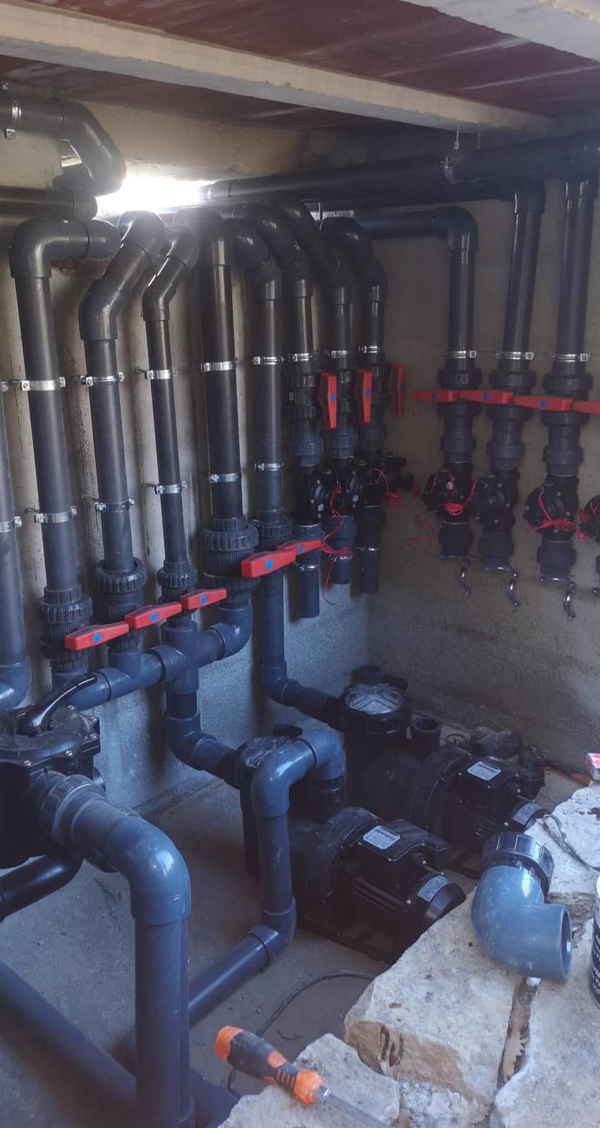 Sala de bombas industrial con tuberías de acero inoxidable, válvulas rojas y grandes bombas sobre un suelo de hormigón.