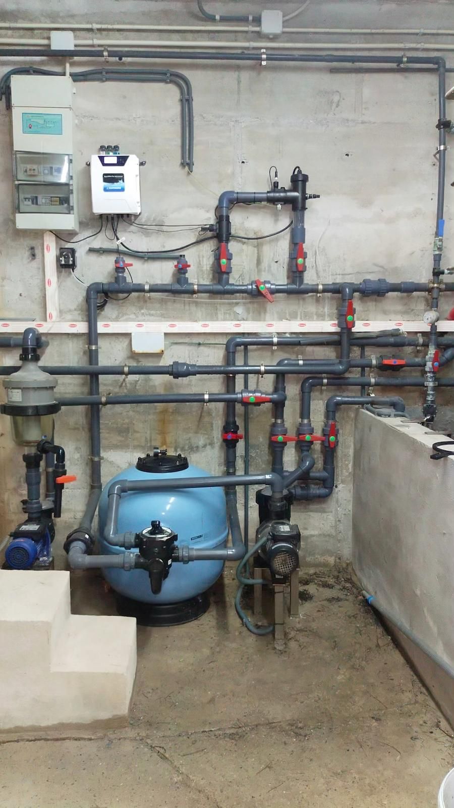 Cuarto de servicio en el sótano con tanque de agua azul, tuberías, válvulas y contadores eléctricos en una pared de hormigón.