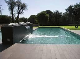Piscina moderna en el patio trasero con pared de agua negra, agua turquesa y terraza de piedra bajo los árboles.