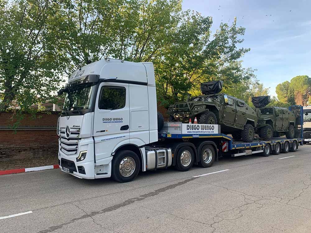 Un camión blanco transporta un vehículo militar en un remolque.