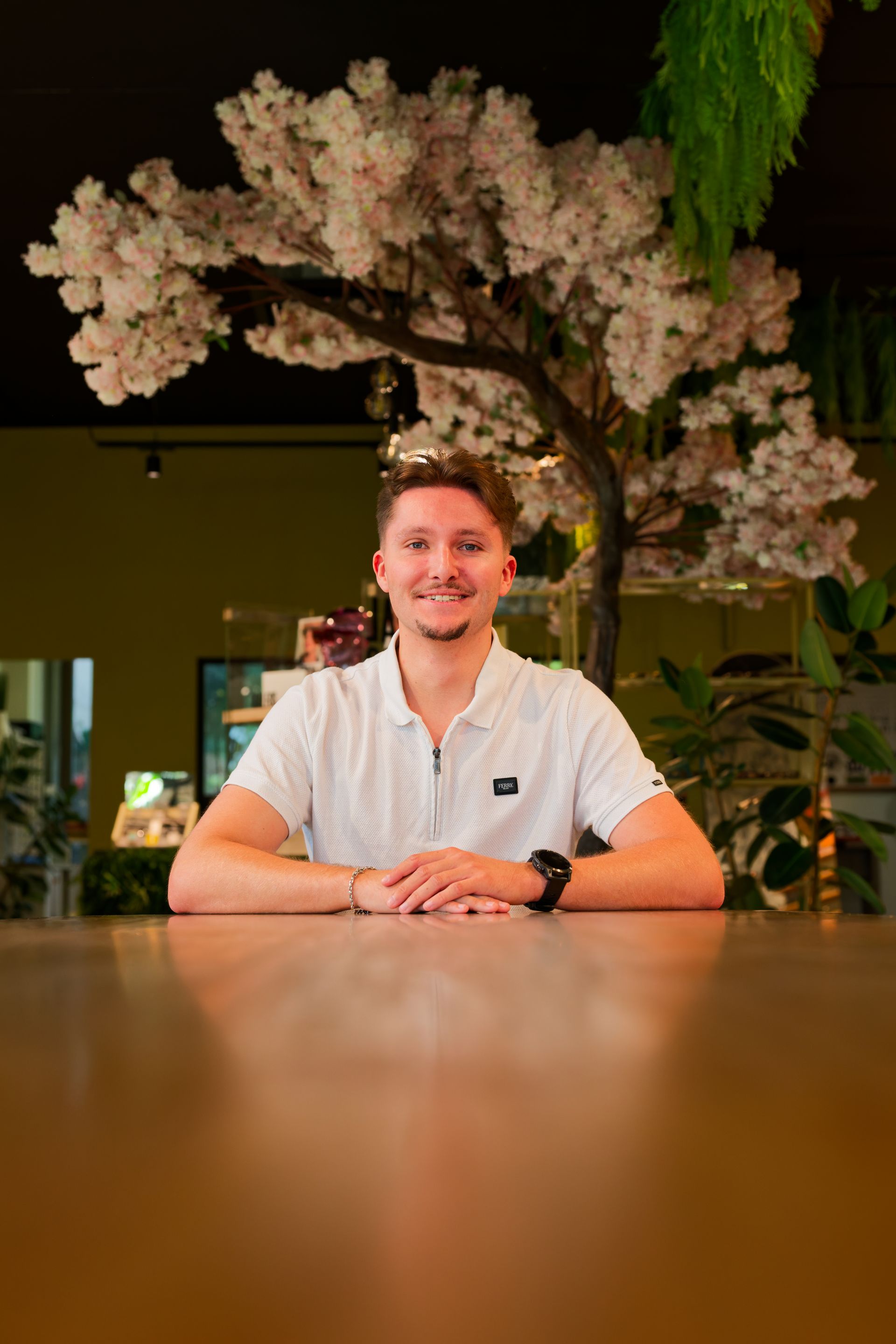 Une personne assise à une table devant un arbre en fleurs, souriant dans un cadre intérieur chaleureusement éclairé