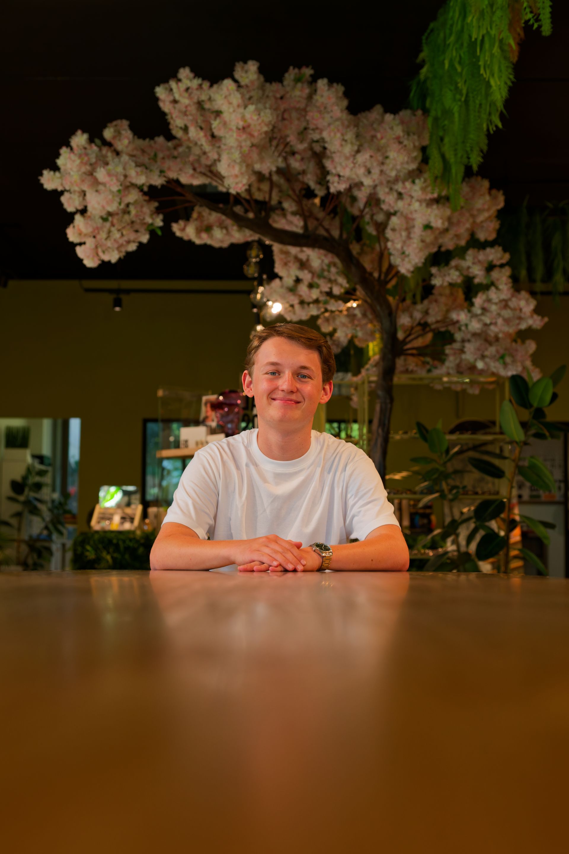 Une personne assise à une table à l'intérieur, souriante, avec un arbre en fleurs derrière elle.