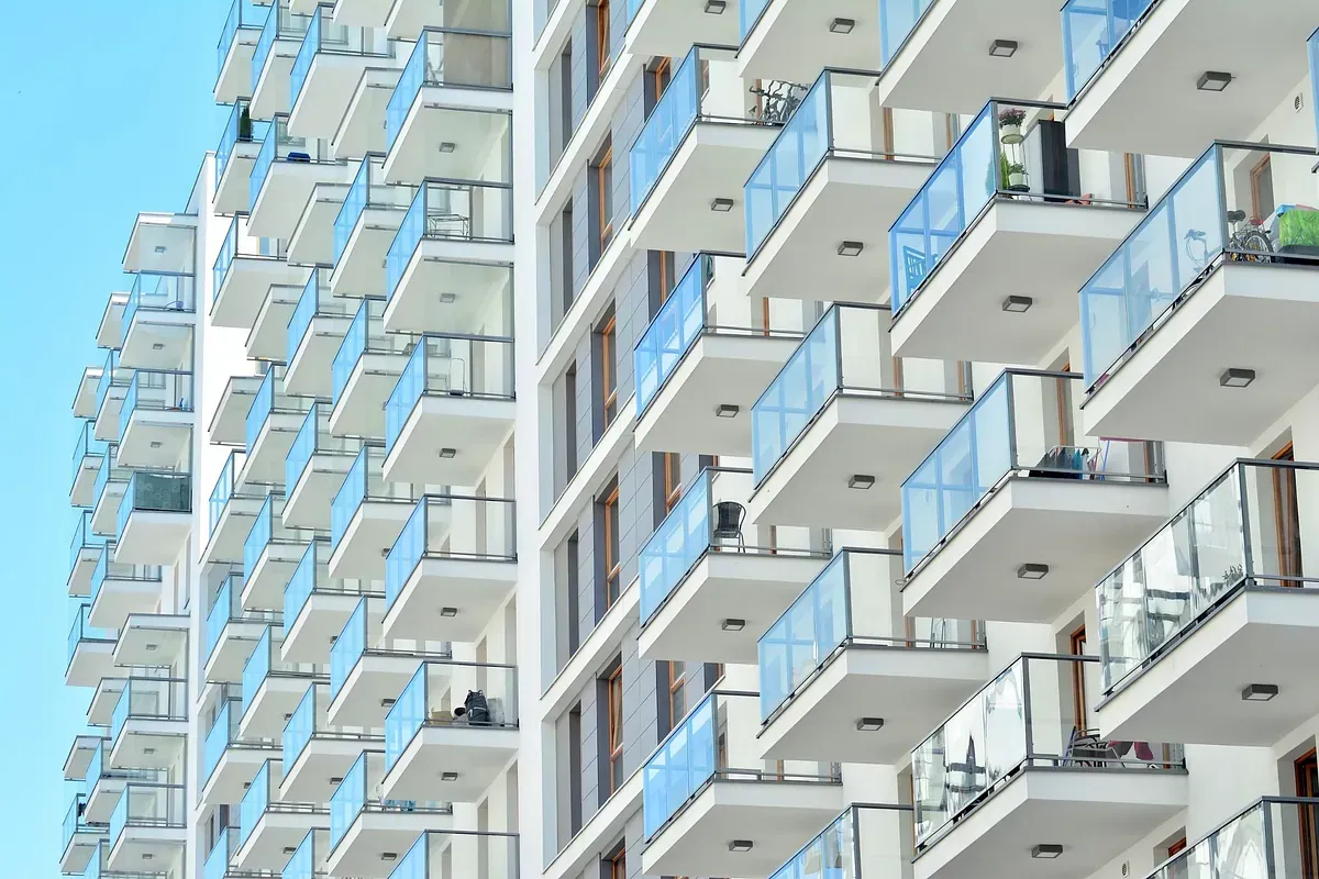 Edificio moderno, de color blanco, de gran altura, con numerosos balcones, barandillas de cristal azul y un cielo azul claro.