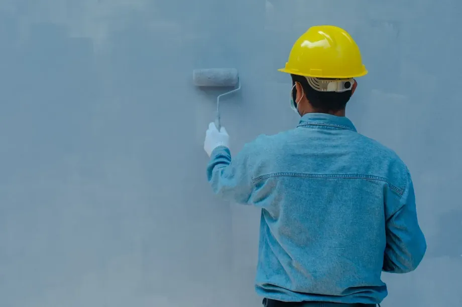 Persona con camisa vaquera y casco amarillo pintando una pared gris con un rodillo.
