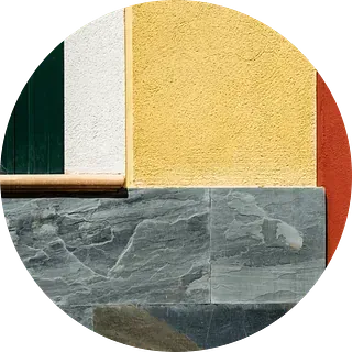 Imagen redonda: Pared exterior con secciones texturizadas de color amarillo y rojo, marco de ventana blanco y base de piedra gris.