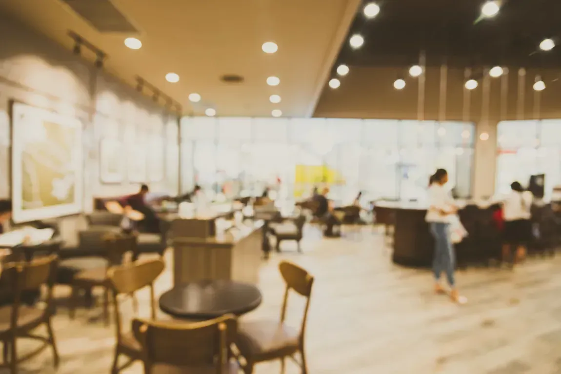 Interior borroso de una cafetería, con mesas, sillas y clientes; tonos cálidos.