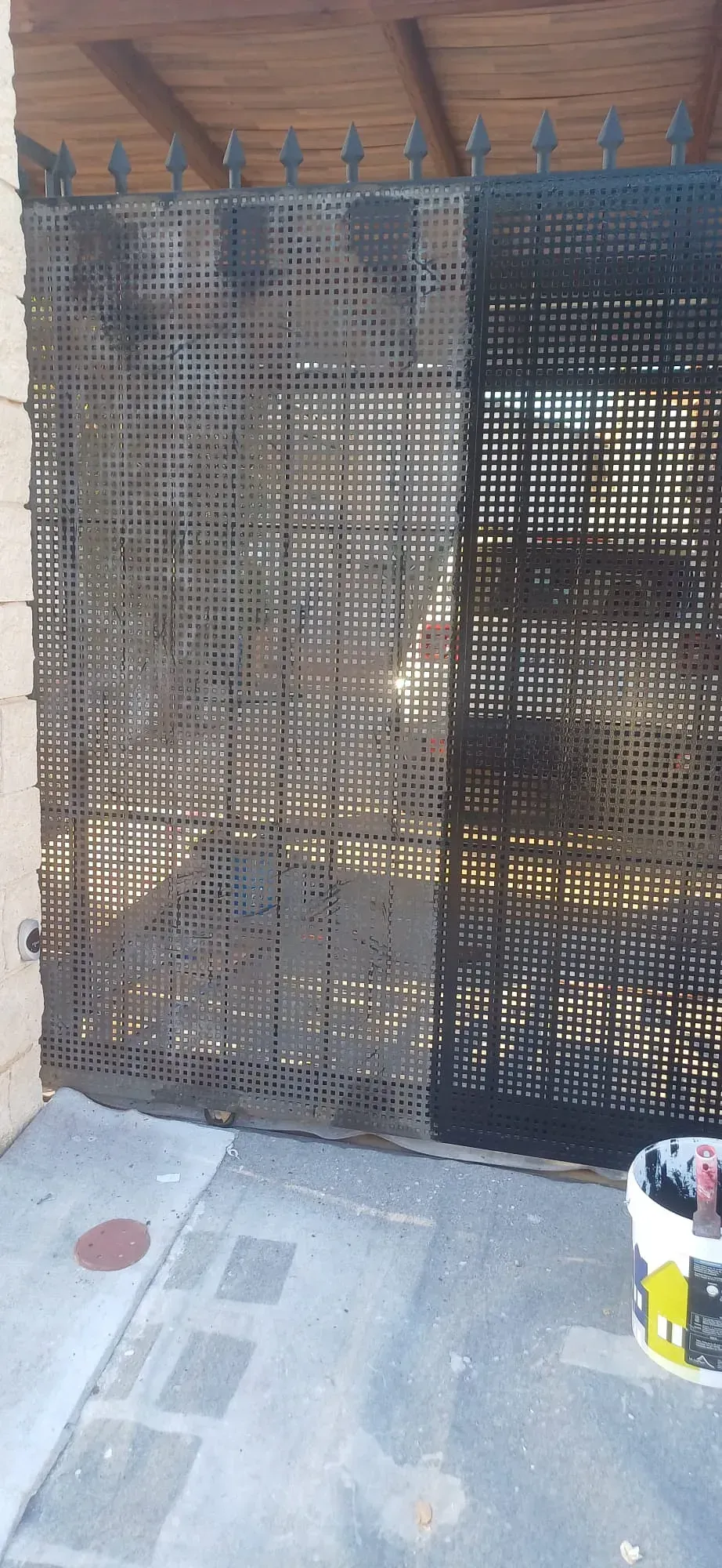 Una valla metálica con una malla oscura, parcialmente pintada. Un cubo de pintura yace en el suelo.