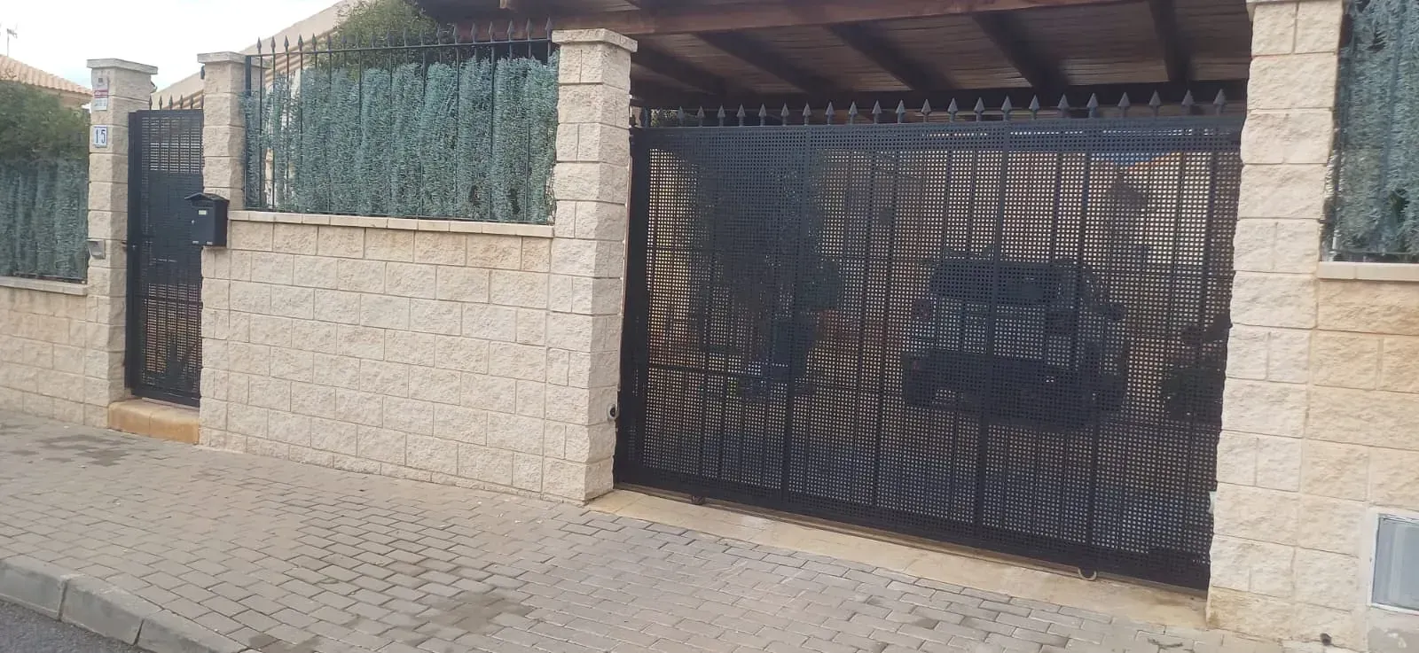 Puerta de metal negro con patrón texturizado, colocada en una pared de piedra beige con setos en la parte superior.