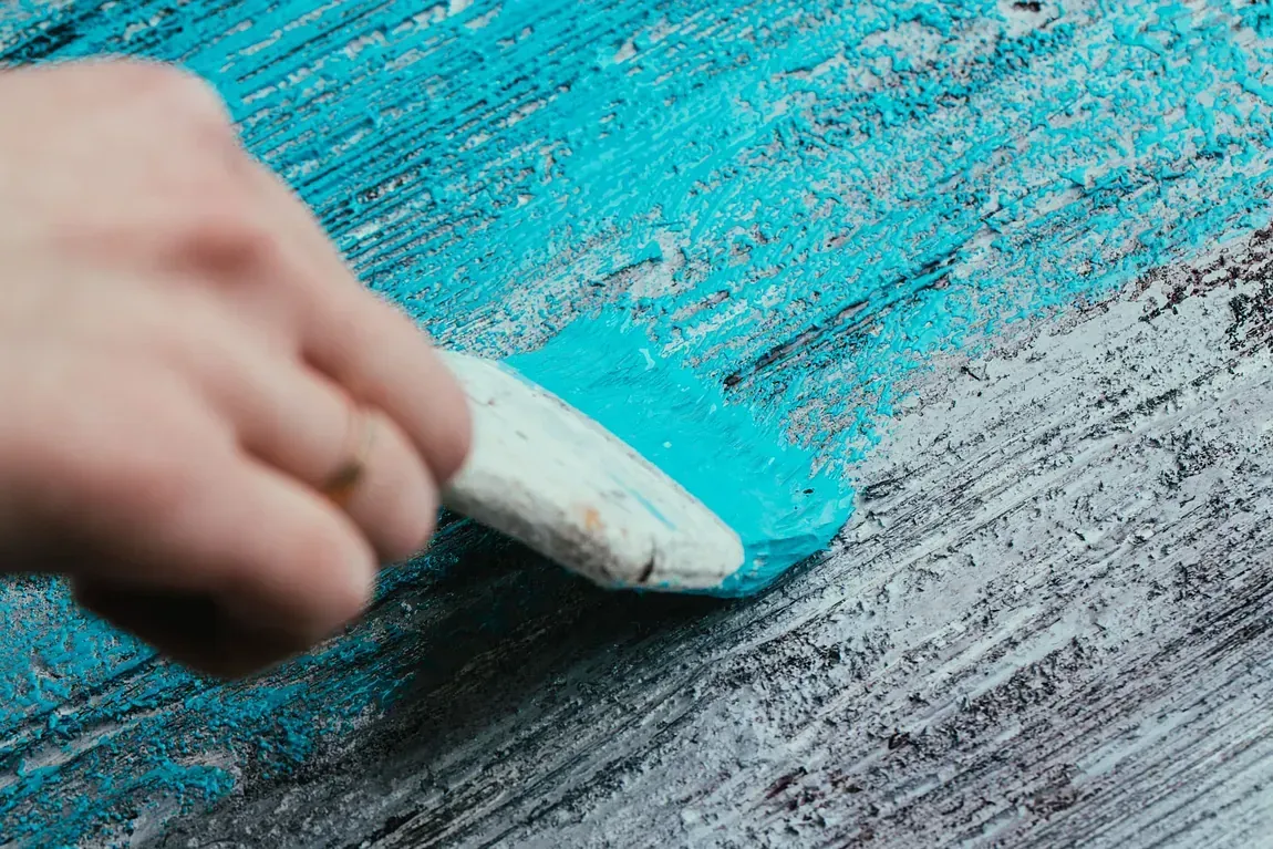 Mano que utiliza un pincel para aplicar pintura turquesa sobre una superficie gris desgastada.