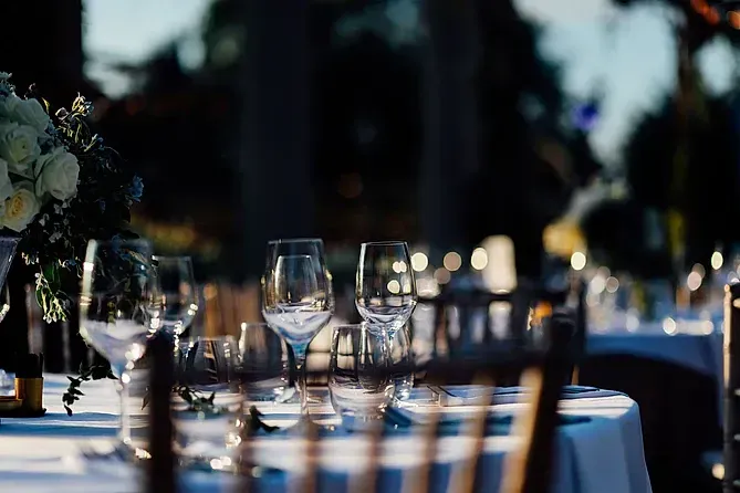Mesa de comedor al aire libre con vasos, flores y sillas, iluminación tenue, ambiente nocturno.