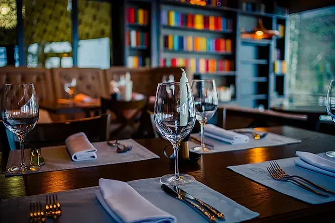 Mesa de restaurante puesta con copas de vino, servilletas y cubiertos, frente a una estantería.