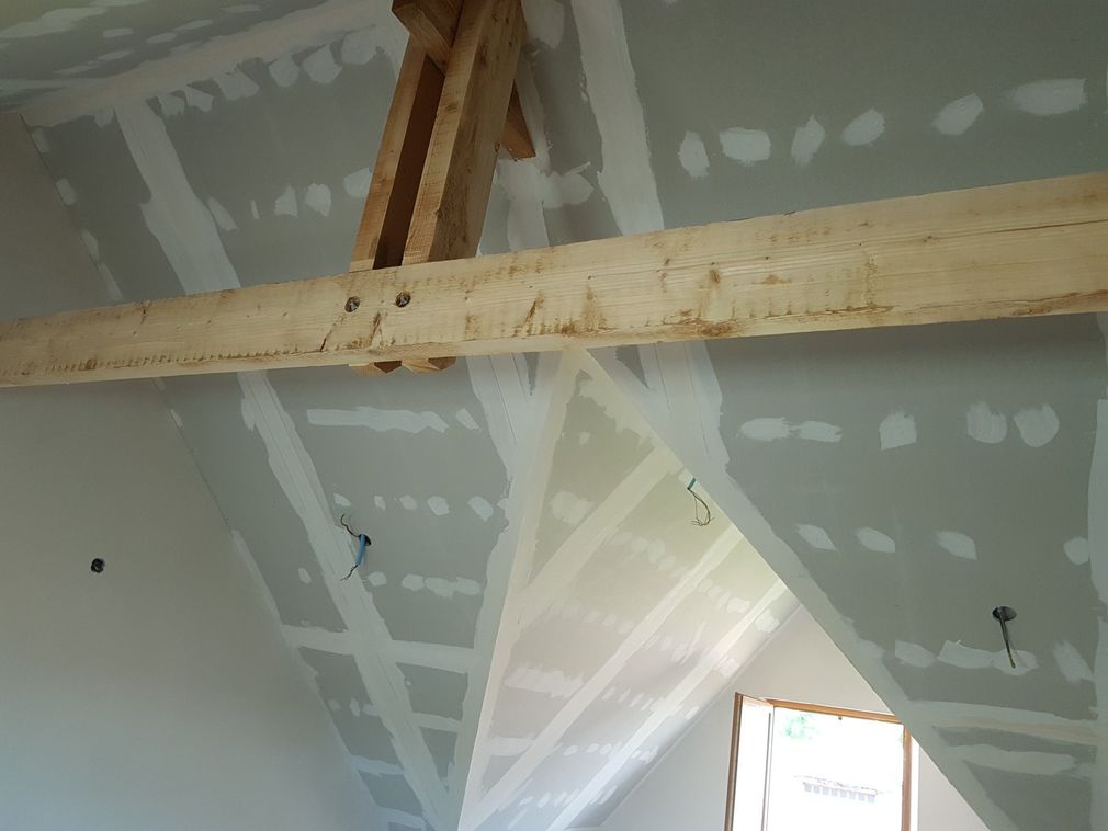 Plafond de grenier non aménagé avec poutres en bois apparentes, joints de plaques de plâtre et une ouverture de fenêtre.