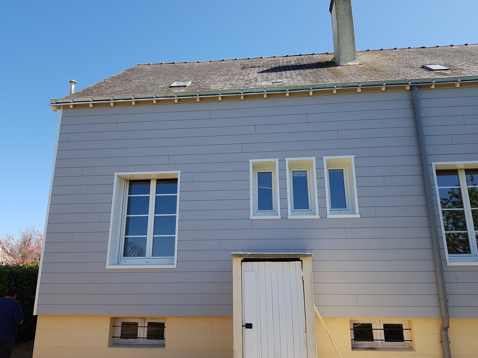 Façade d'une maison grise aux boiseries blanches, fenêtres, porte blanche et toit de tuiles sous un ciel bleu