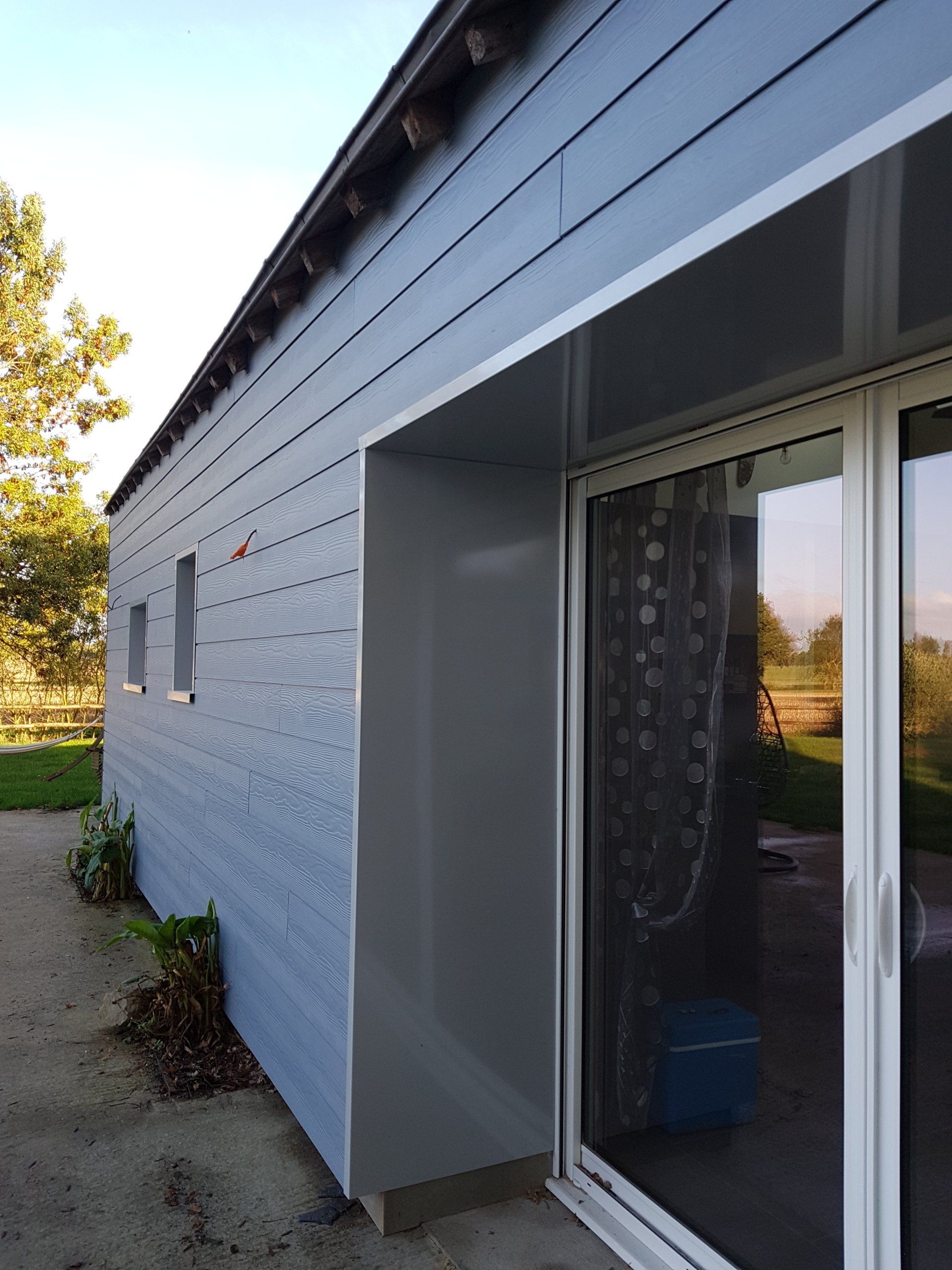 Aménagement intérieur Extérieur de maison moderne avec bardage gris, fenêtres latérales étroites et porte-fenêtre coulissante donnant sur un patio.
