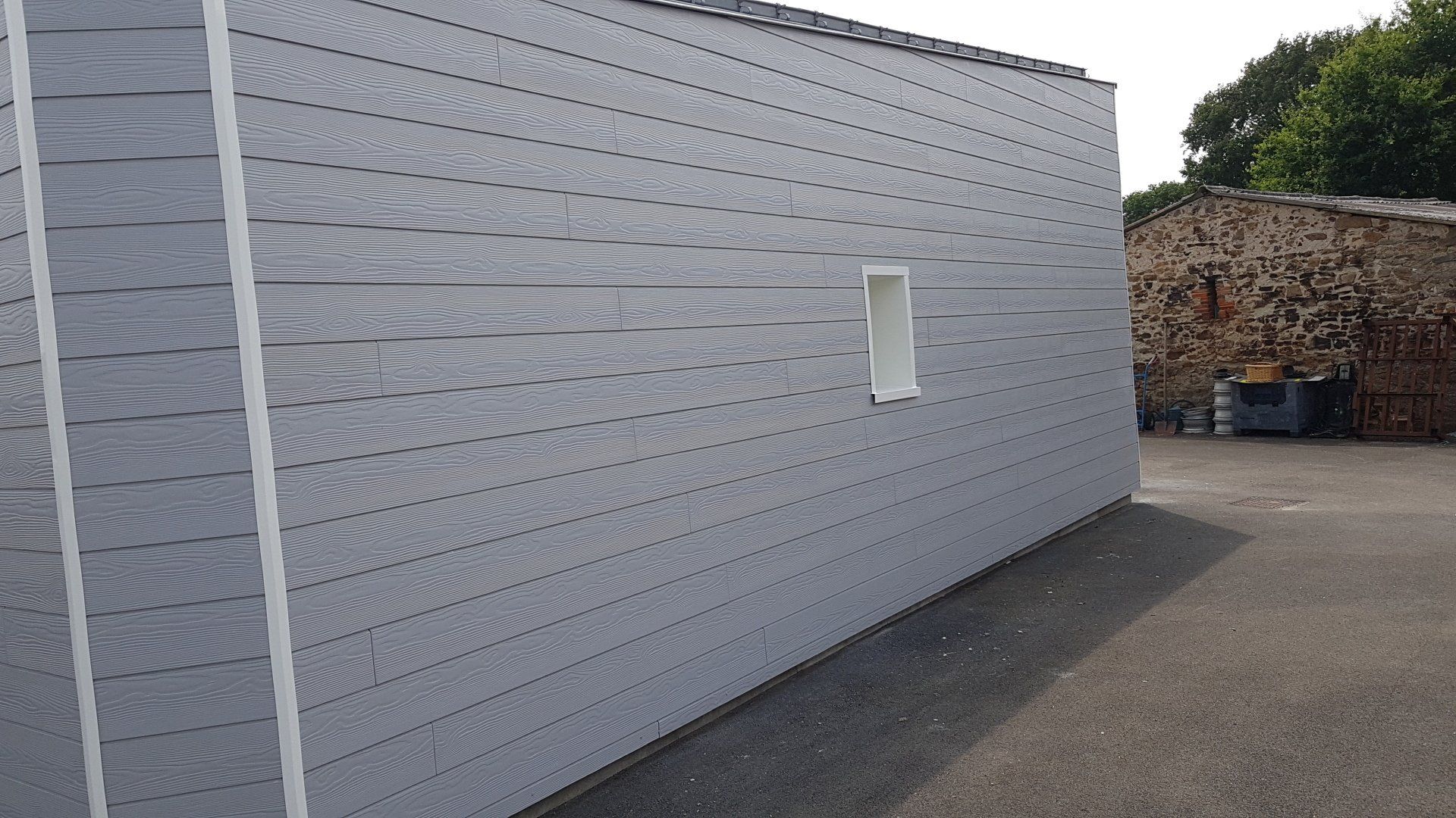 Mur blanc d'un bâtiment industriel avec une petite fenêtre et une zone pavée à côté.
